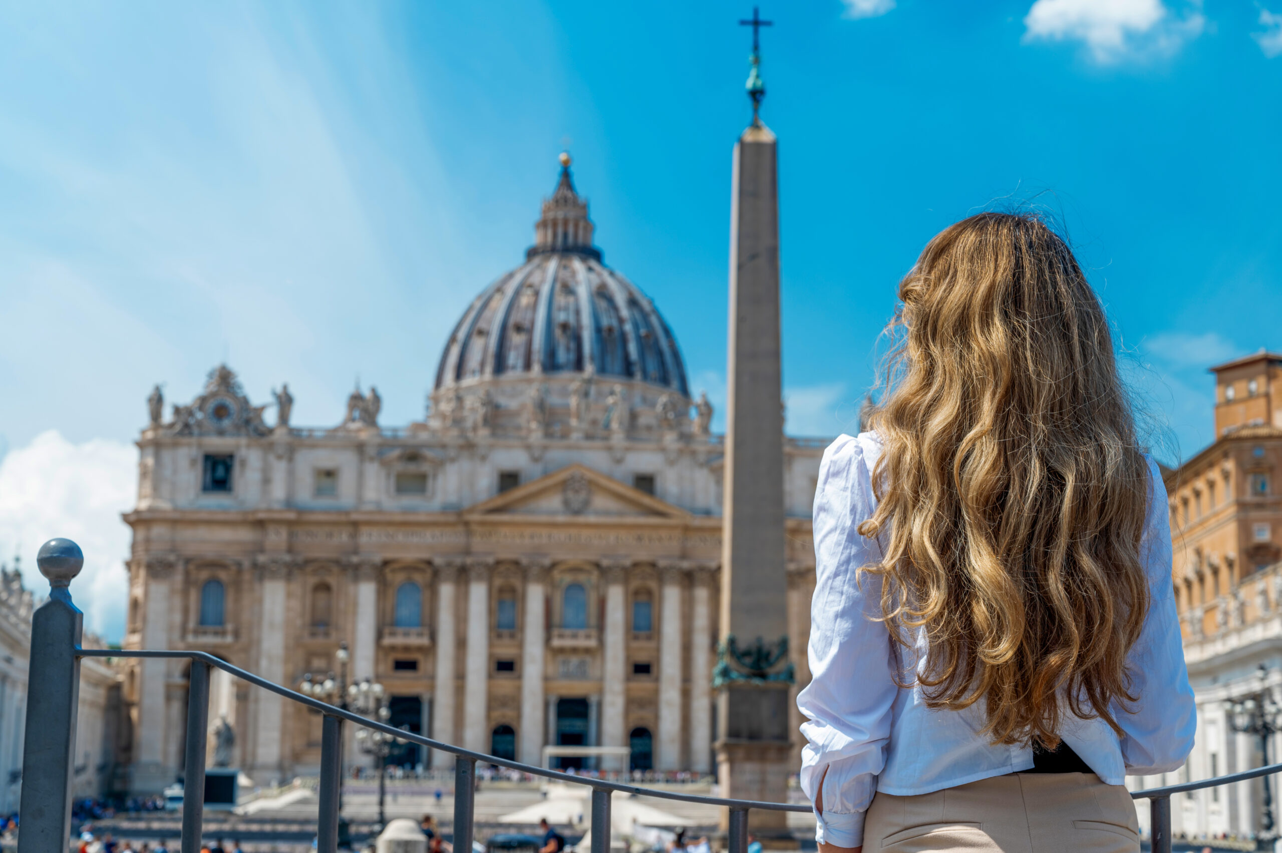 View of the blonde woman from behind in Vatican City. Saint Peter Square with multiple tourists, Saint Peter Basilica and the Vatican Obelisk on the background